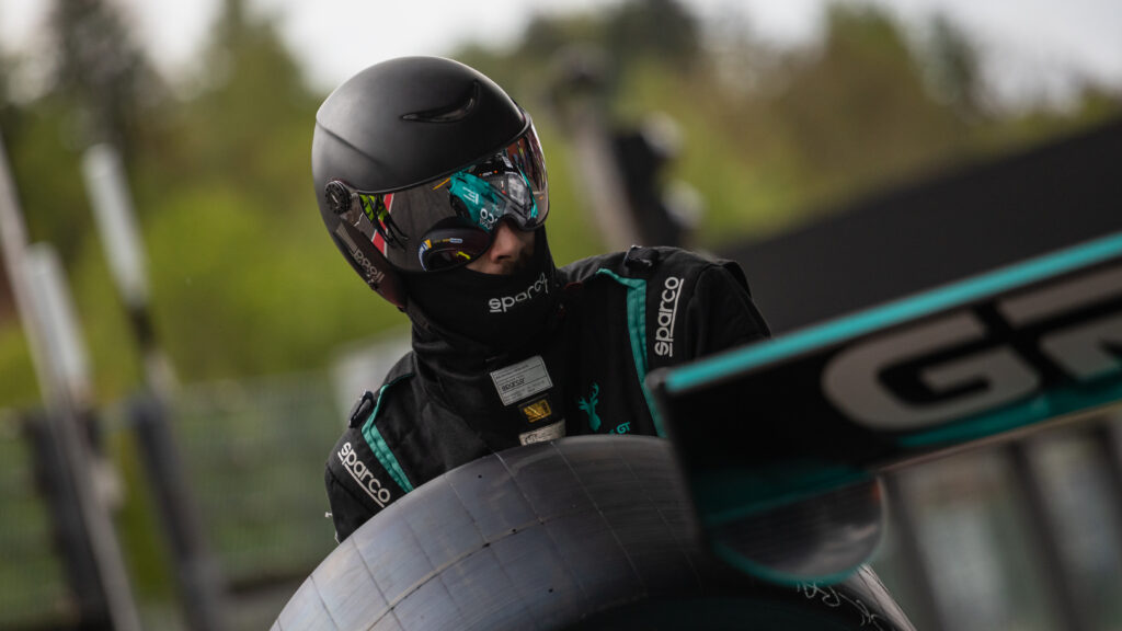 Pitcrew member holding a tire, during the Pitstop of his car. He wears a helmet with  a reflecting visor where you can see a blue car a McLaren from Greyston GT